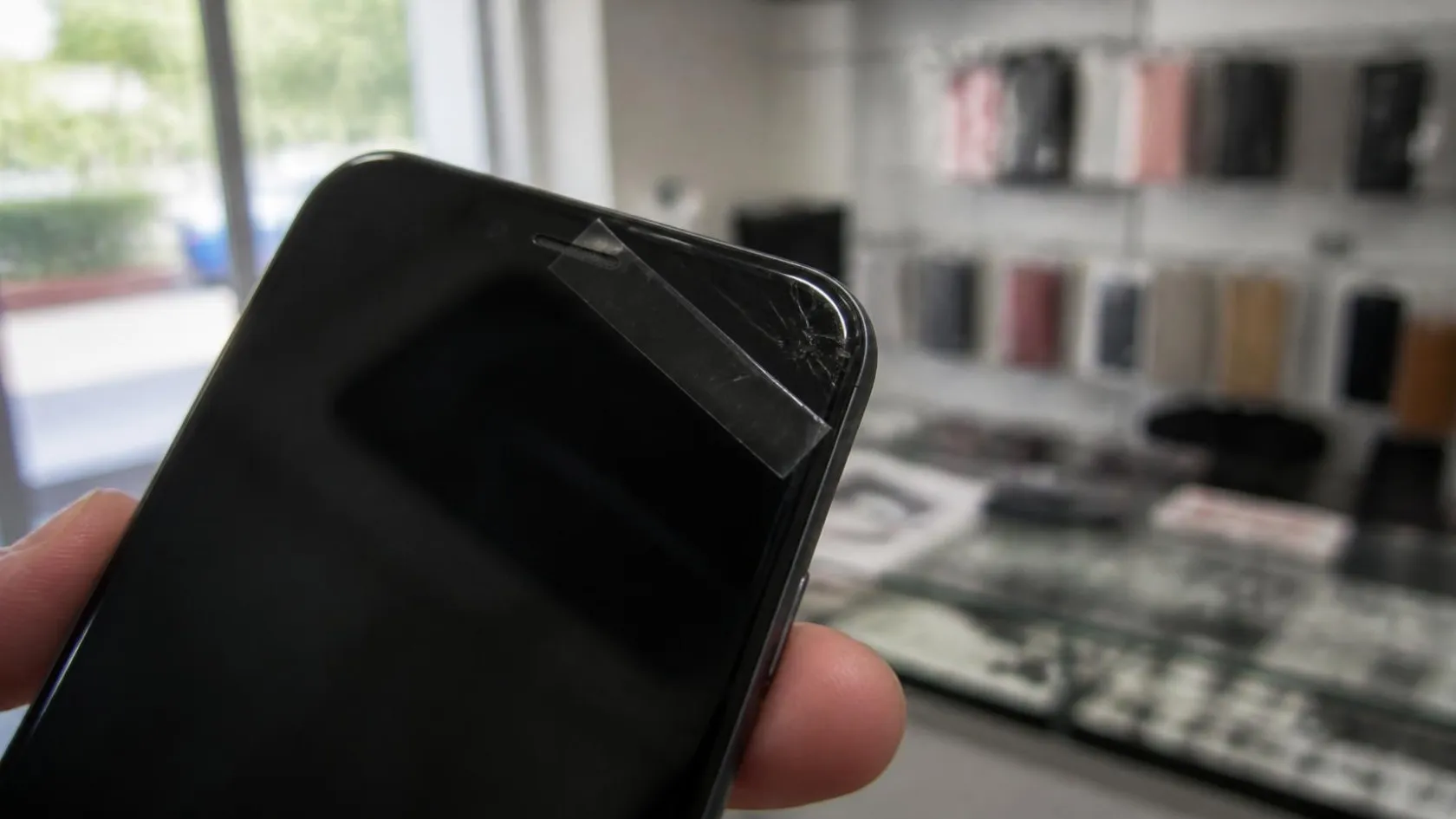 Hand hält ein Smartphone, ein Streifen Klebeband liegt über der abgesplitterten Glasecke mit Haarrissen