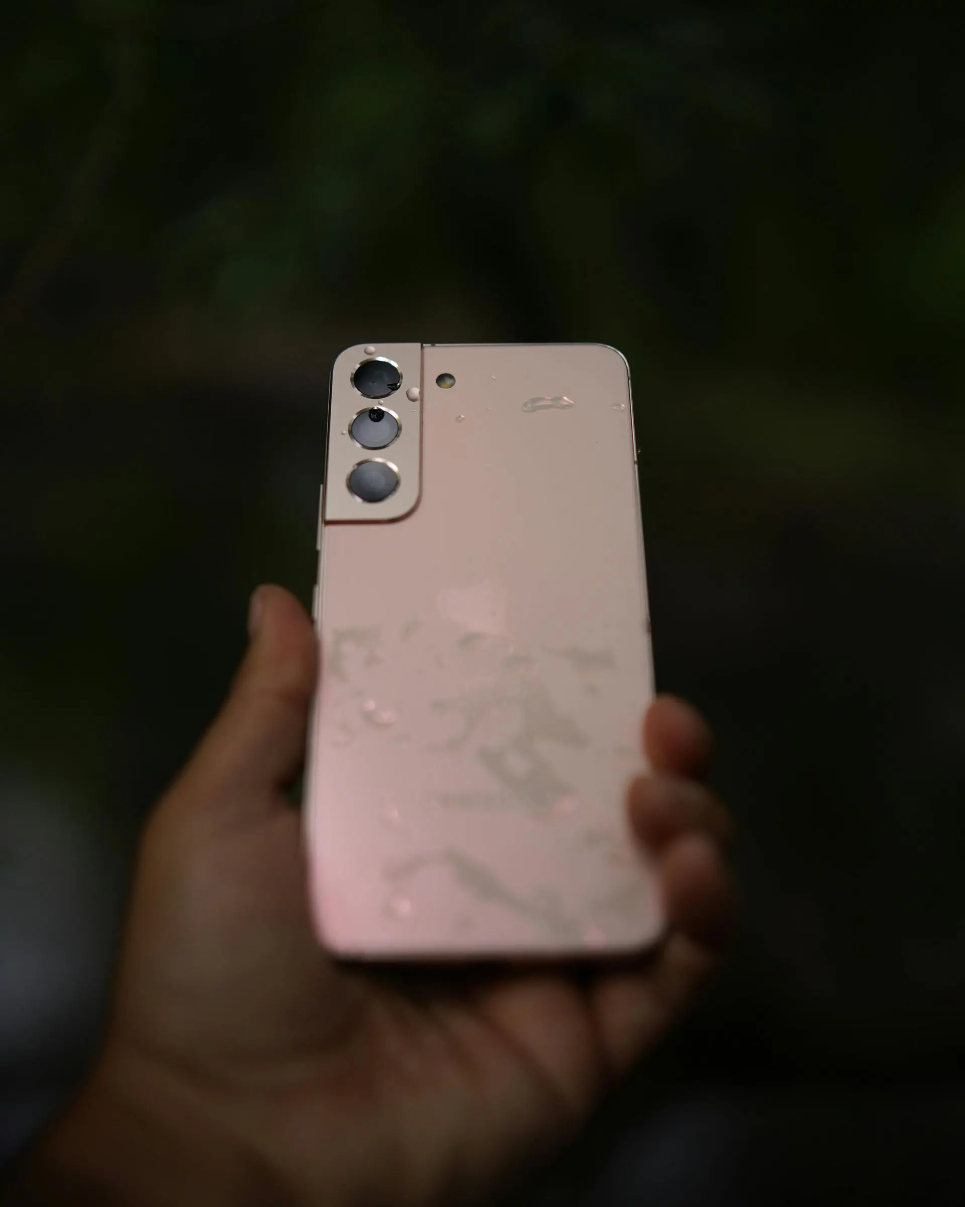 Eine Hand hält ein pinkfarbenes Samsung-Handy mit drei Kameras, das mit Wassertropfen benetzt ist, vor einem dunklen, verschwommenen Hintergrund.