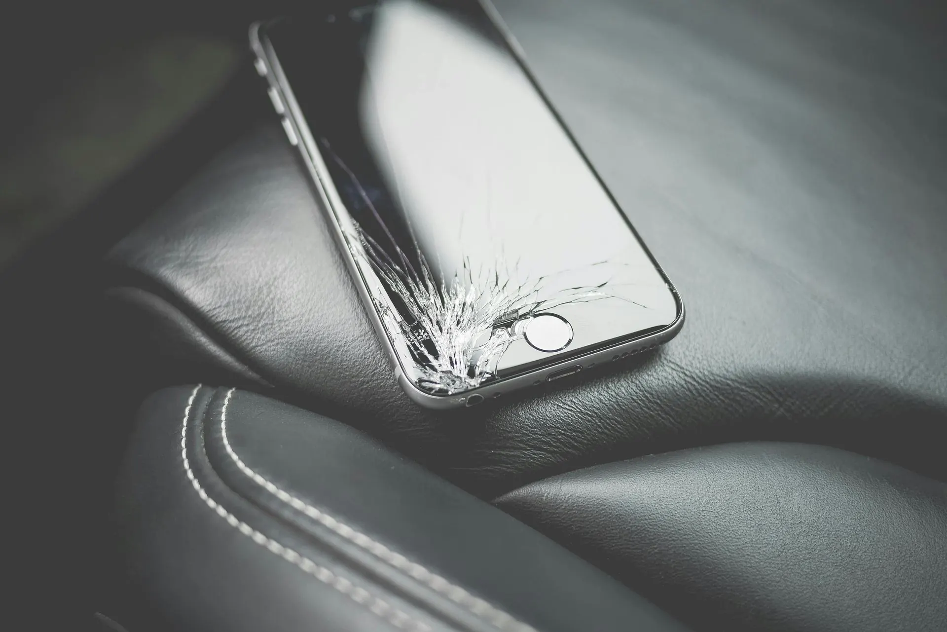 Ein gesprungener Smartphone-Bildschirm liegt auf einem schwarzen Ledersitz neben einer weiß abgenähten Armlehne.