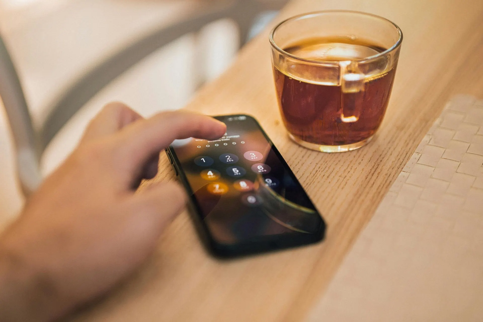 Eine Hand berührt den Bildschirm eines Smartphones neben einem Glas Tee auf einer Holzoberfläche.