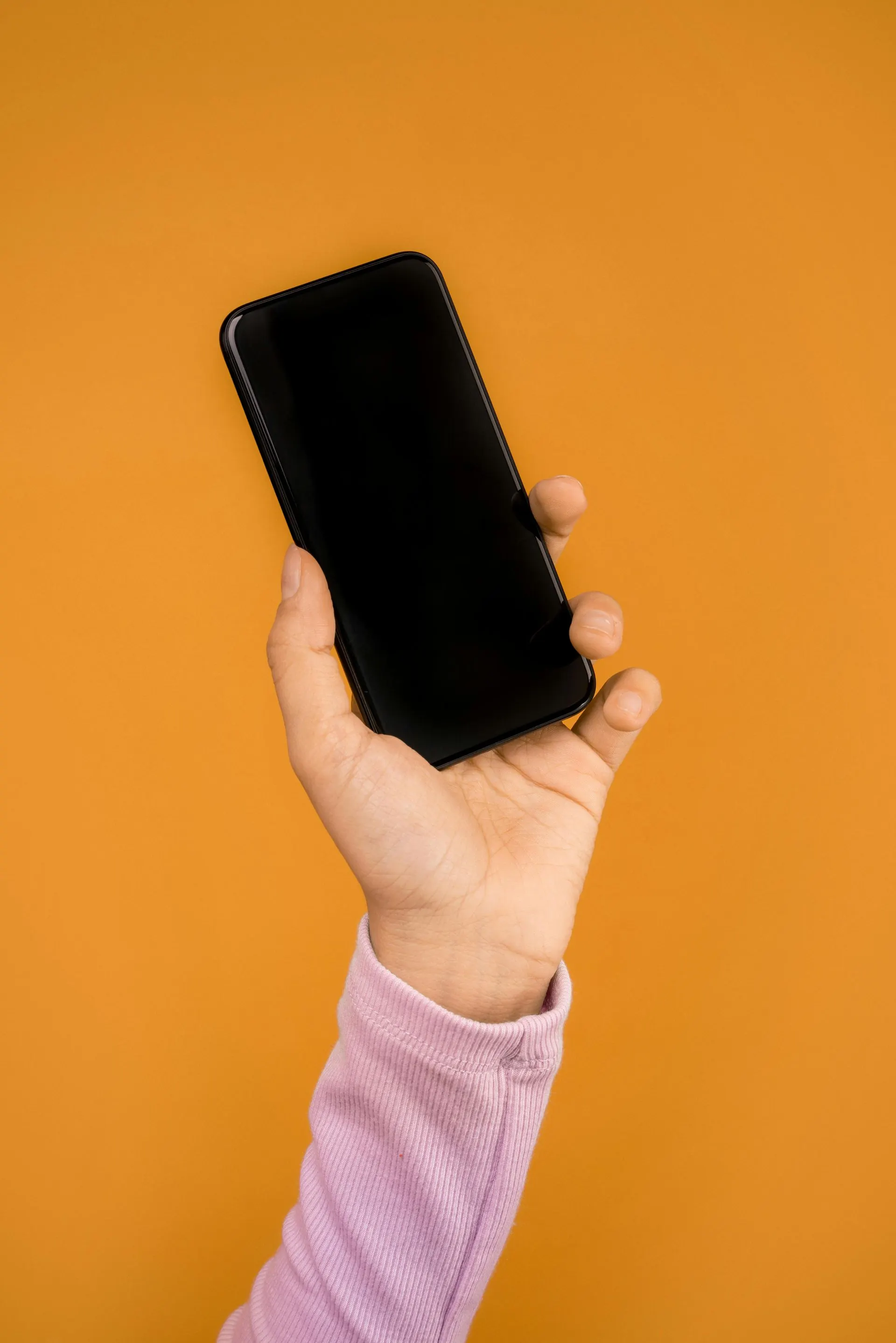 Hand hält ein schwarzes Smartphone mit leerem Bildschirm vor orangefarbenem Hintergrund.