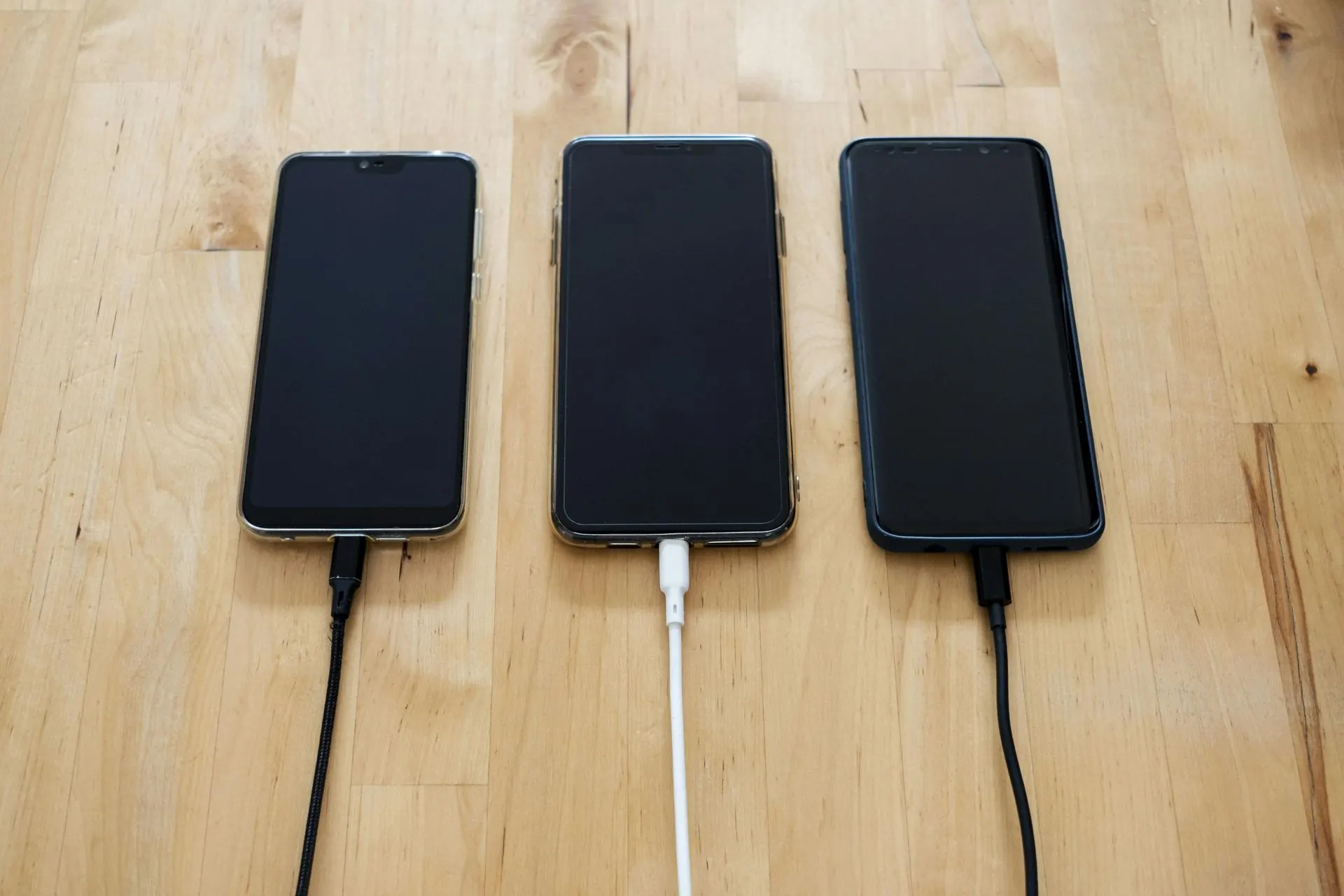 Drei schwarze Smartphones laden auf einer Holzoberfläche, jedes mit einem andersfarbigen Ladekabel.