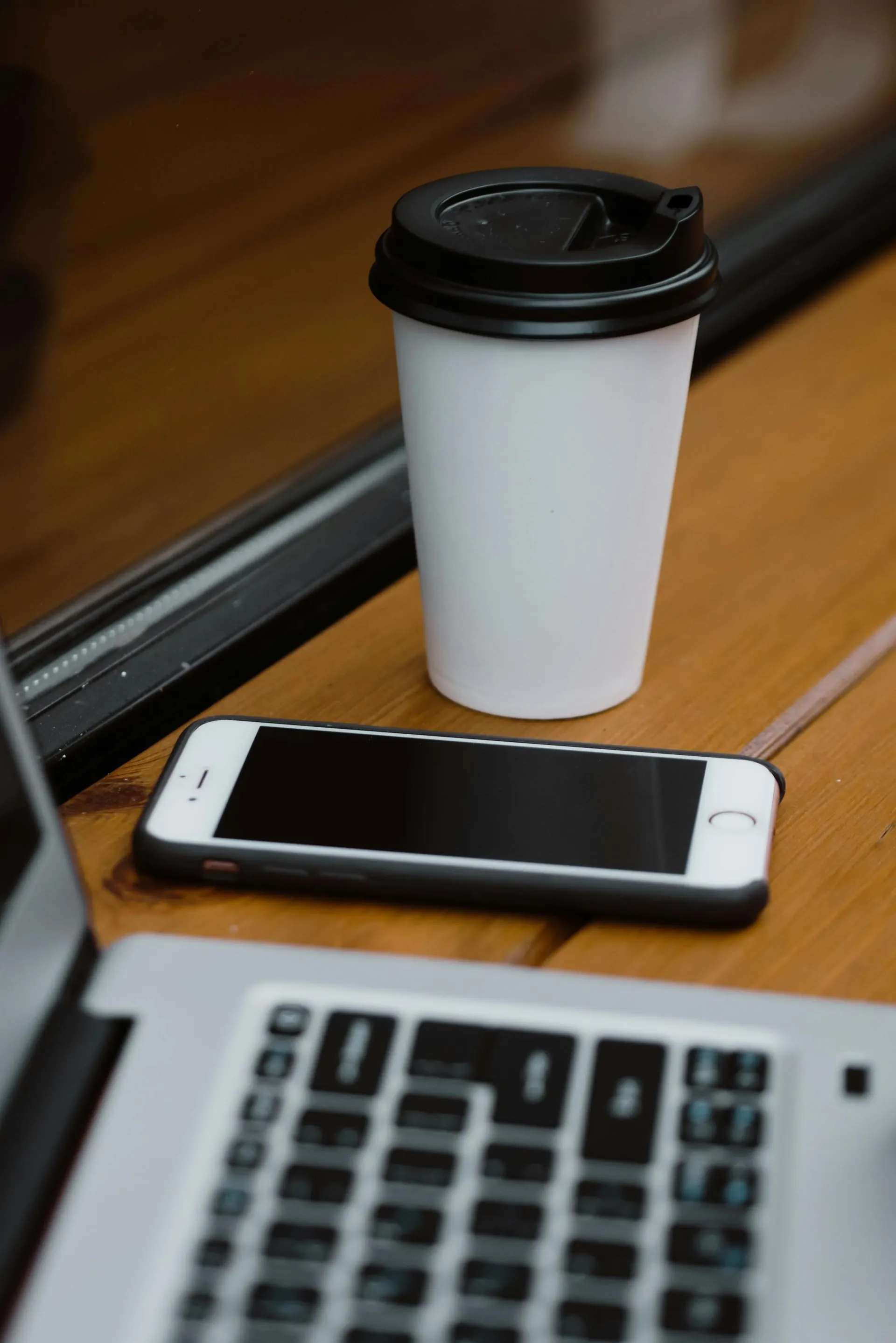 Weiße Kaffeetasse und Smartphone auf einem Holzschreibtisch neben einer Laptop-Tastatur.